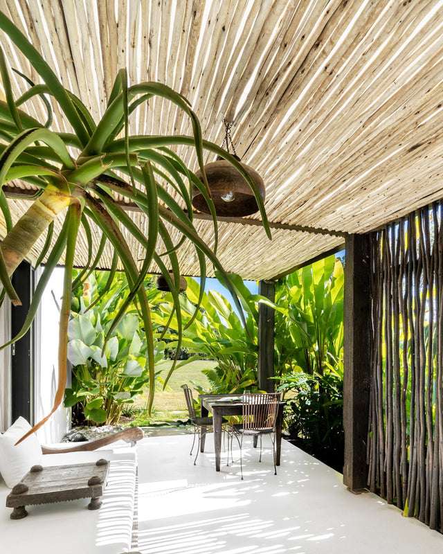 Tropical outdoor dining area under a eucalyptus pole pergola, featuring lush greenery, a rustic wooden dining table with metal chairs, and hanging pendant lighting. The space exudes a relaxed, resort-style ambiance