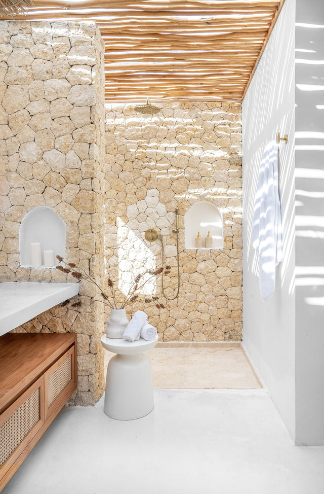 Tranquil outdoor shower with natural stone walls, brass fixtures, timber slatted ceiling, and white rendered details, styled with soft towels and earthy decor for a day spa atmosphere.