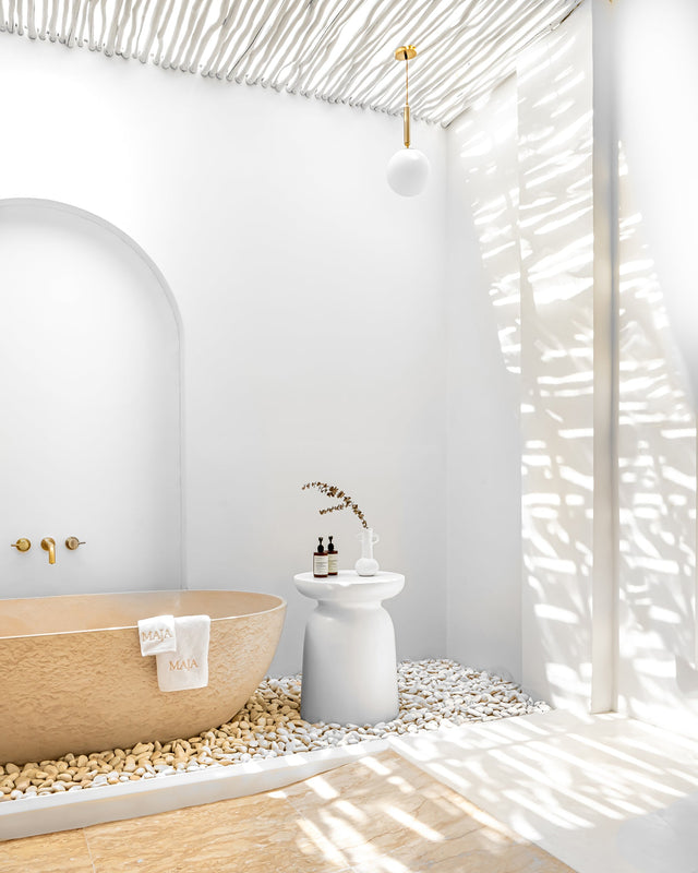 Tranquil outdoor bathroom with a sculptural stone bathtub surrounded by smooth white pebbles, minimalist white side table, and natural eucalyptus pole roof casting dappled light.