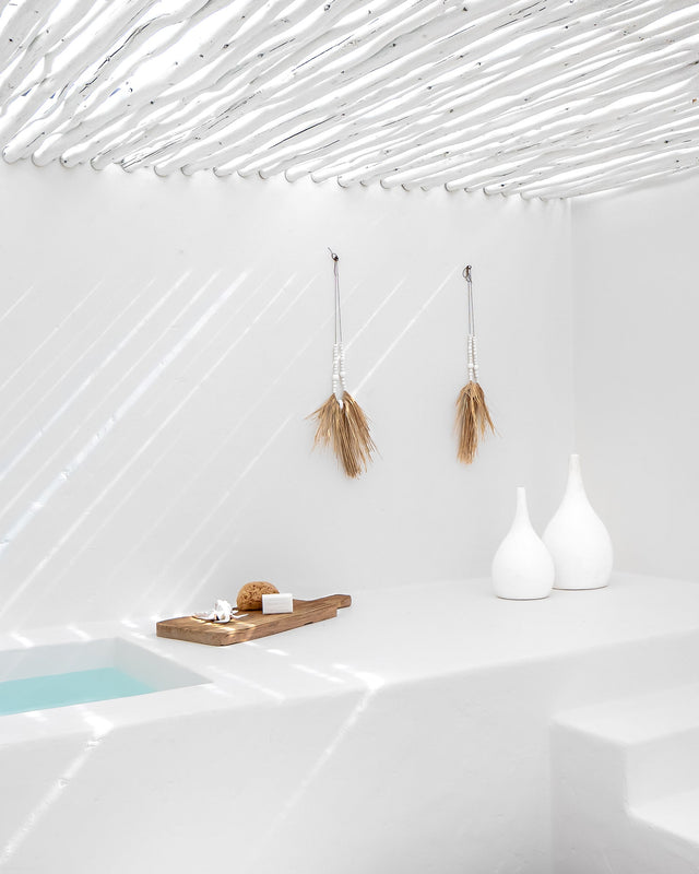 Serene outdoor day spa featuring a white rendered bath, natural timber bath tray, decorative white vases, and pampas grass wall hangings beneath sculptural white ceiling rafters.