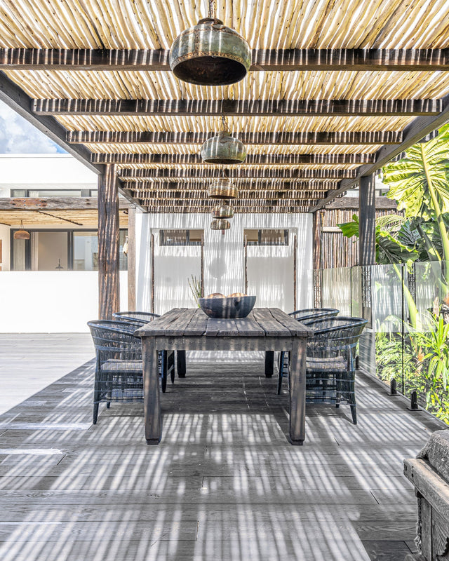 Outdoor dining area featuring a black timber dining table and woven chairs under a rustic eucalyptus pergola, with handcrafted pendant lights and lush tropical surroundings.