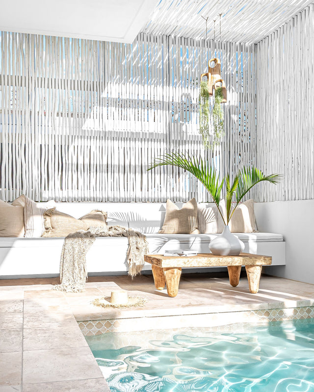 Bright and airy poolside lounge featuring a white built-in sofa with neutral cushions, a rustic wooden coffee table, and a tropical plant in a ceramic vase. Eucalpytus pergola and natural textures create a relaxed, coastal atmosphere