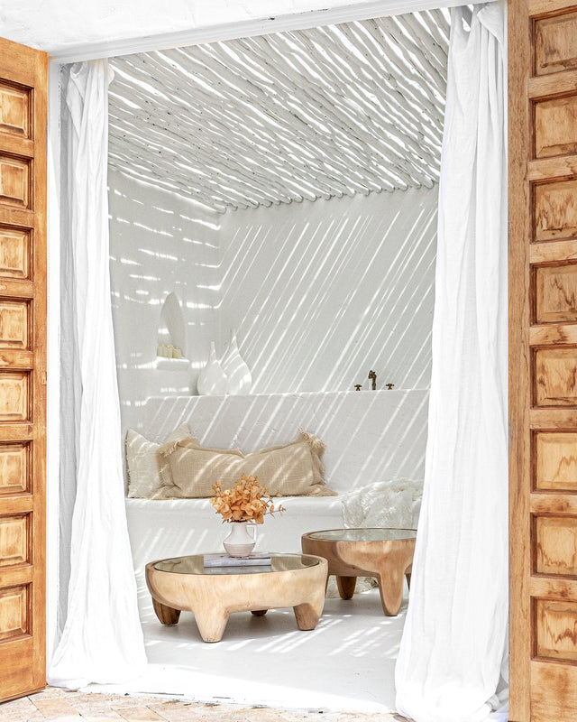 Bright coastal-style lounge featuring curved glass-top wooden coffee tables, textured neutral cushions, and slatted overhead shadows creating a tranquil, sun-dappled effect.