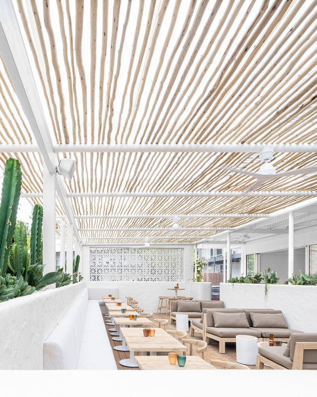 Bright and airy outdoor seating area under a white bamboo pergola, featuring natural timber tables, beige lounge seating, and a cactus planter for a relaxed, resort-style atmosphere